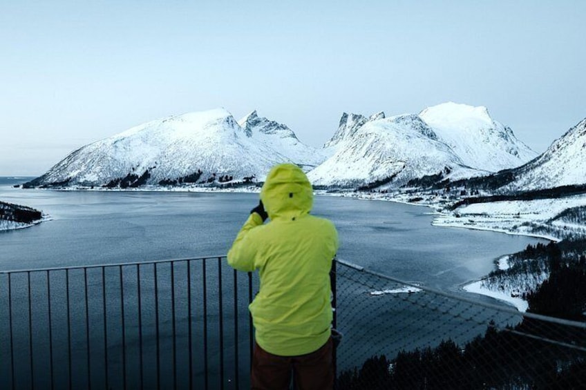 A person in a yellow jacket taking a photo of a snow-covered landscape with mountains and a frozen lake. 3