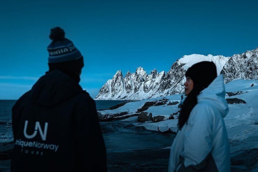 A person in a black jacket with a logo, another in a white jacket, and a snowy mountainous landscape in the background. 4