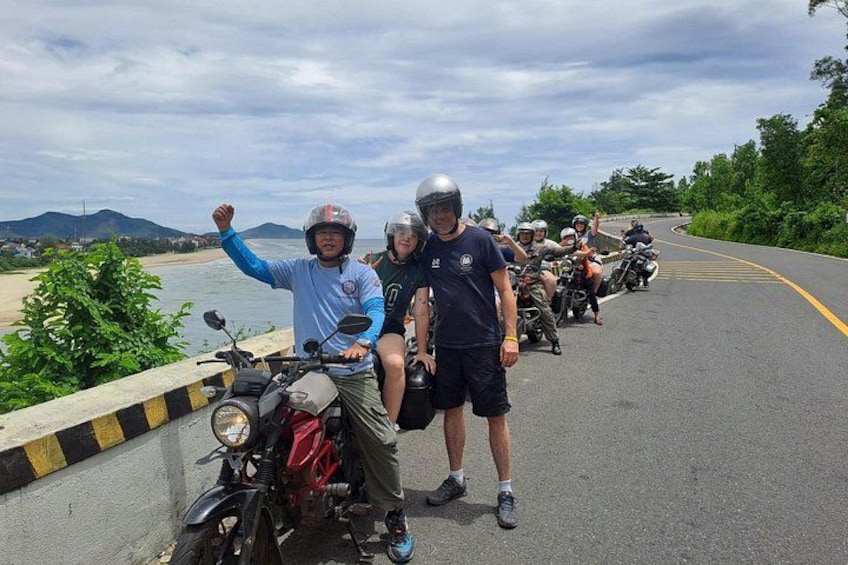 A group of motorcyclists on a bridge with a coastal landscape in the background. 1