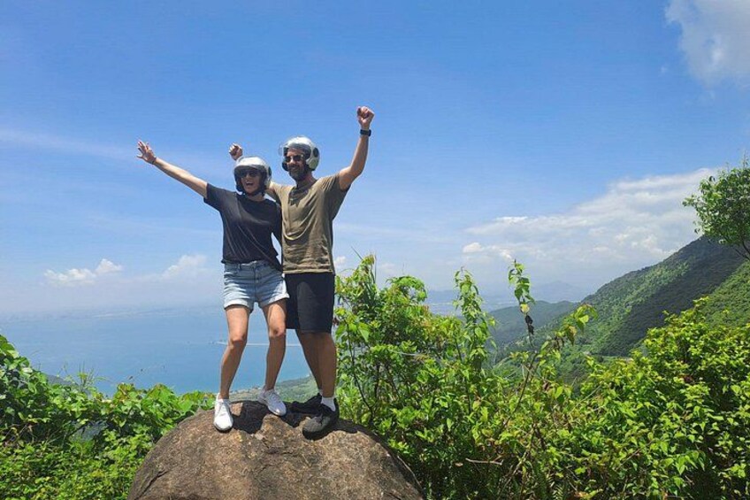 Two people standing on a rock with arms raised, surrounded by greenery and a clear sky. 2