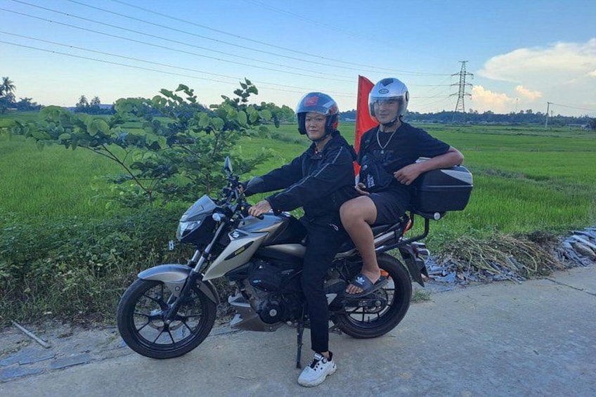 Two people on a motorcycle with a red flag, a rural road, and green fields in the background. 4