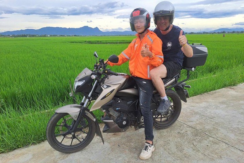 A motorcycle with two people, one giving a thumbs up, parked on a road with green fields and mountains in the background. 5