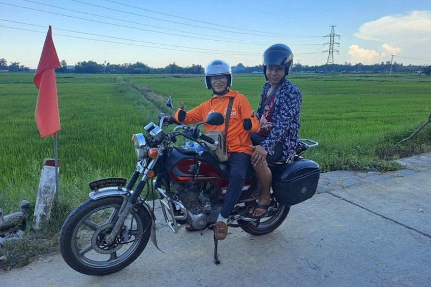 A motorcycle with two riders on a rural road, surrounded by green fields. 3