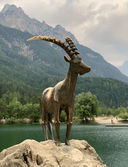 A bronze statue of a goat on a rock by a lake with mountains in the background. 3