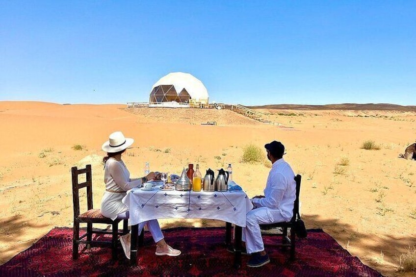 Two people dining at a table in a desert with a large dome structure in the background. 1
