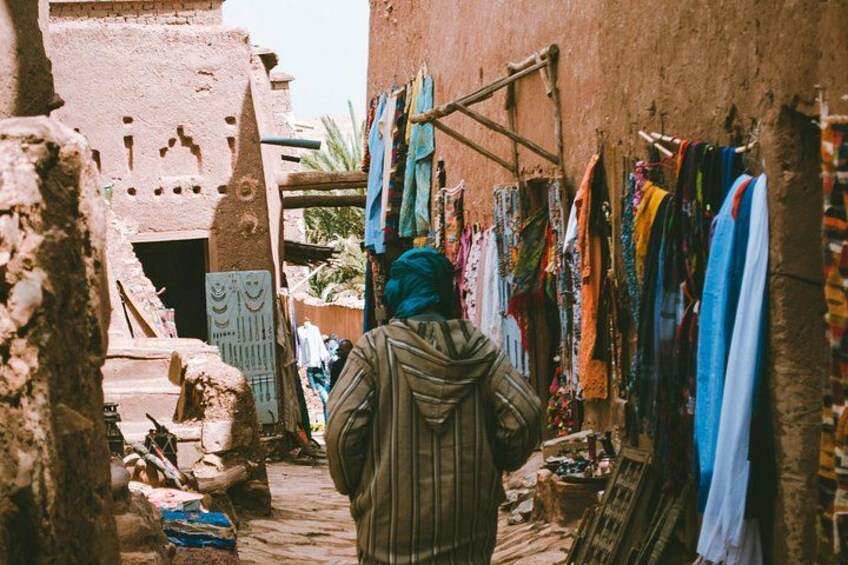 A narrow alley with colorful textiles hanging on racks, a person in traditional attire, and a building with a distinctive facade. 2