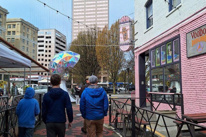 A group of people walking on a sidewalk, a pink building with a sign, and a large colorful sculpture. 1