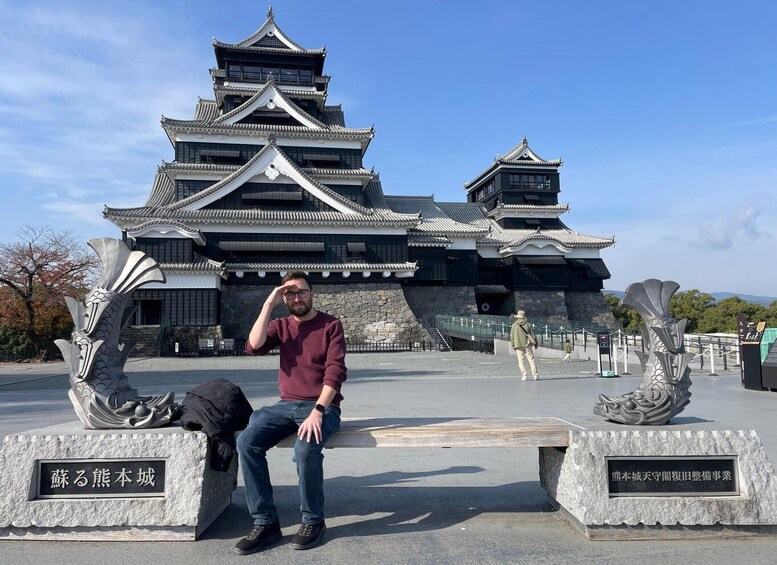 A man sitting on a bench in front of a traditional Japanese castle with two stone sculptures on either side. 4
