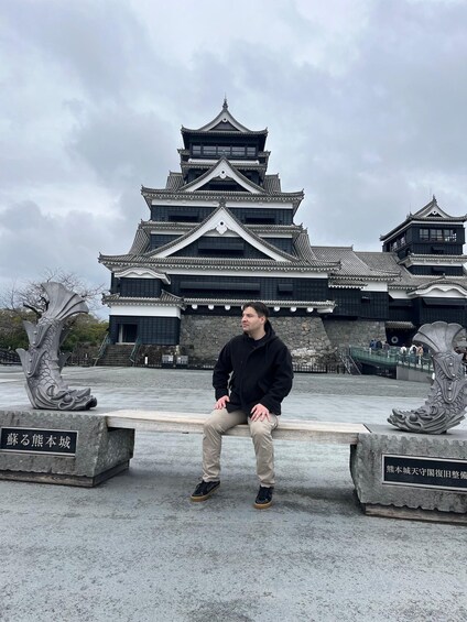 A person sitting on a bench in front of a traditional Japanese castle with ornate architecture and two dragon sculptures. 2