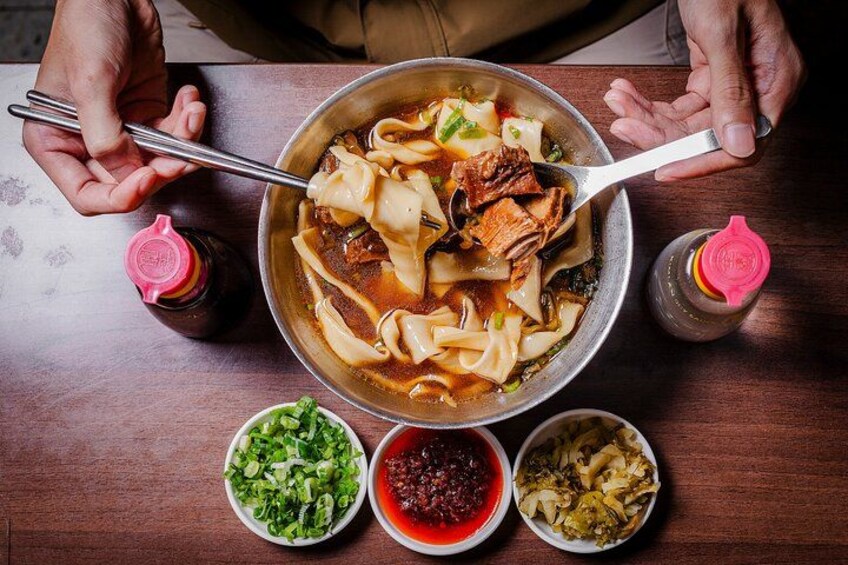 Un bol de soupe contenant des nouilles et de la viande, accompagné de petits bols de condiments et de légumes, le tout posé sur une table en bois. 2