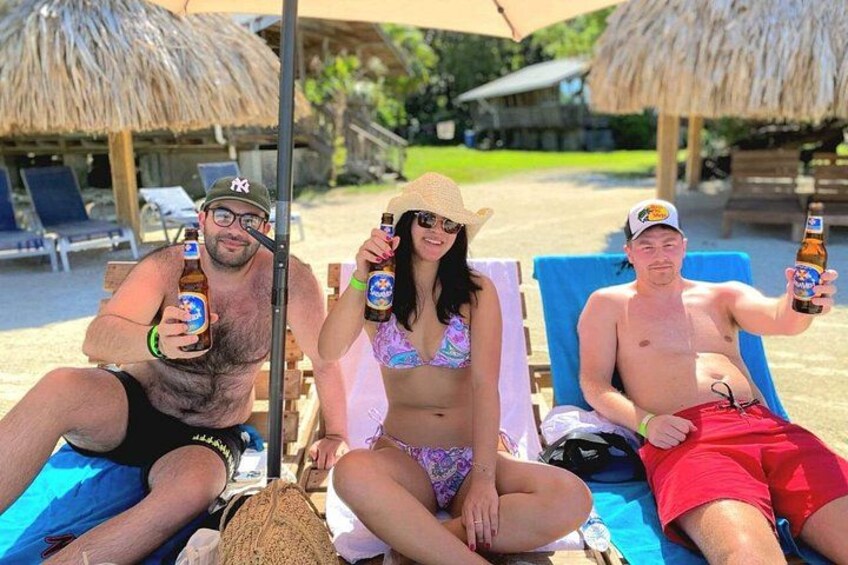 Drie personen in strandstoelen met drankjes, een rieten parasol en een rieten constructie op de achtergrond. 1