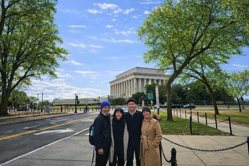 A group of people standing in front of a large building with columns, a tree-lined street, and a clear sky. 2