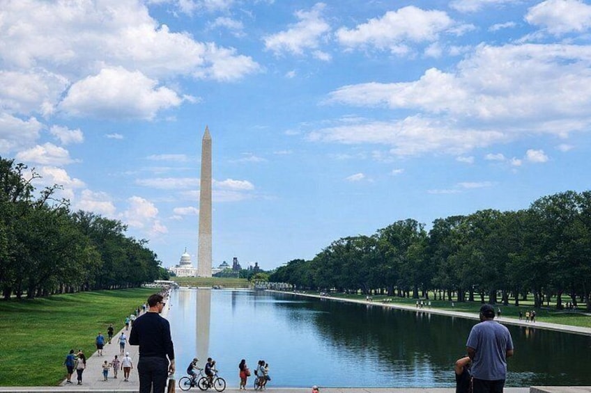 The Washington Monument, a large obelisk, surrounded by a park with a reflecting pool, trees, and people walking and biking. 1