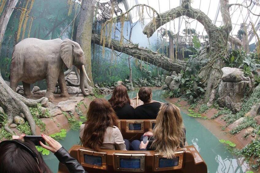 A group of people on a wooden boat ride through a jungle-themed exhibit with an elephant and a river. 1