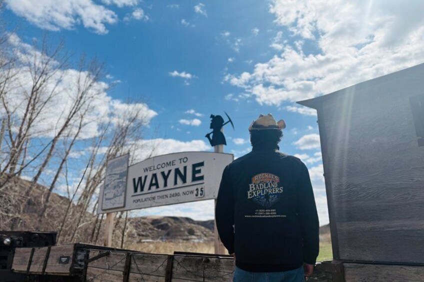 A person standing in front of a 'Welcome to Wayne' sign with a 'Badlands Explorers' jacket, a statue, and a clear sky. 5