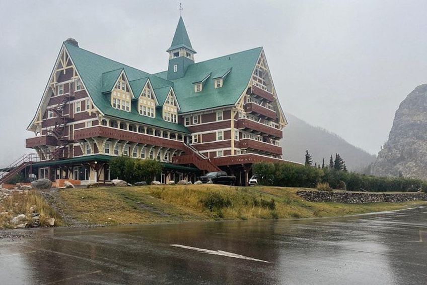A large, multi-story building with a steeply pitched roof and a tower, surrounded by a grassy area and a road. 3