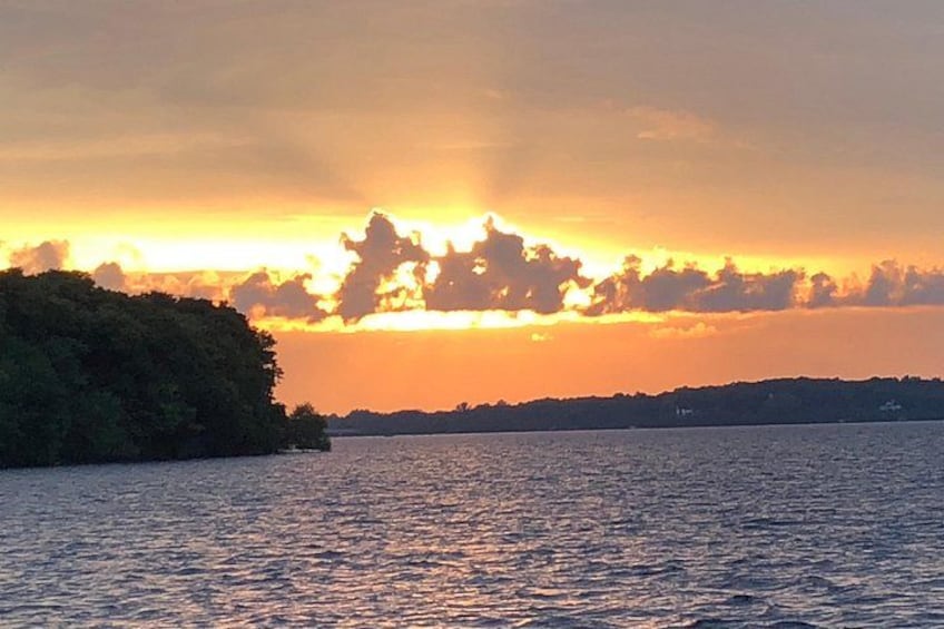 Een zonsondergang boven het water, met een silhouet van land aan de linkerkant en een verre kustlijn aan de rechterkant. 2