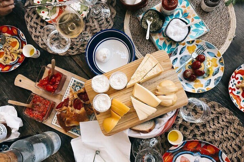 Variedad de quesos, embutidos y condimentos sobre una tabla de madera, rodeados de platos de comida y copas de vino sobre una mesa. 1