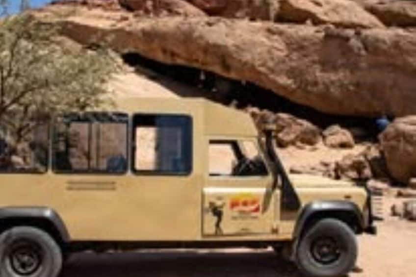 A beige off-road vehicle parked in a desert with large rocks and sparse vegetation. 5