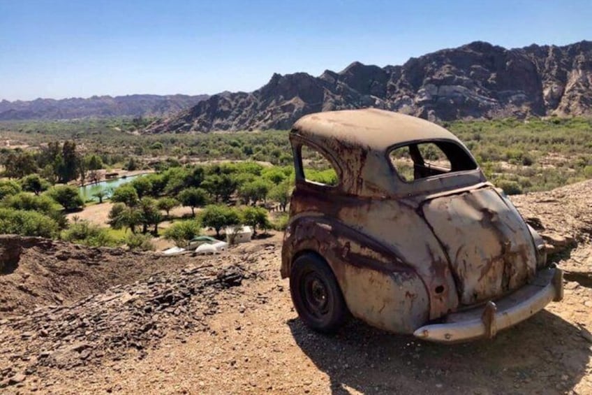 A rusted vintage car parked on a dirt road with a mountainous landscape in the background. 1