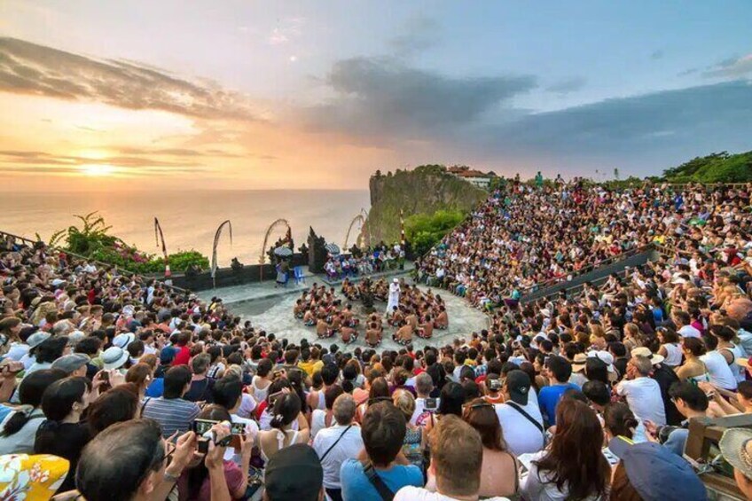 A large crowd at a coastal event with a stage and seating area overlooking the sea. 1