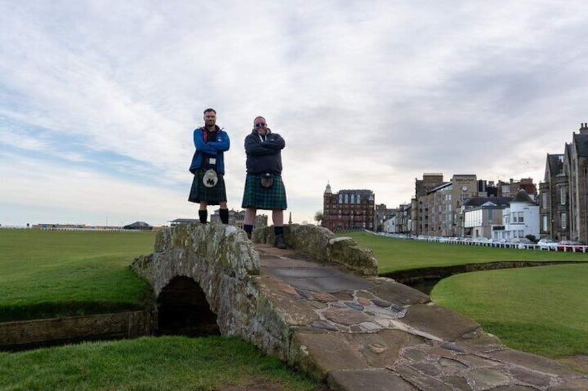 Two men in kilts standing on a stone bridge over a grassy area with buildings in the background. 4
