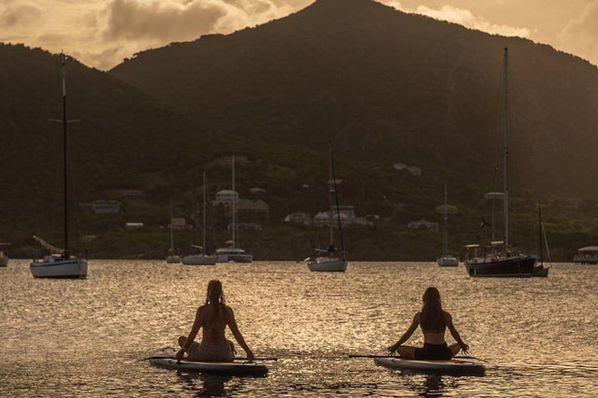 Two people meditating on surfboards in calm waters with sailboats and a mountainous backdrop. 1