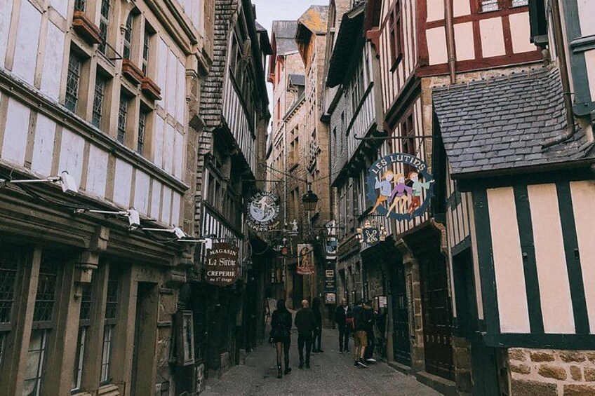 A narrow medieval street with timber-framed buildings, a clock, and a sign for 'Les Loutres.' 3