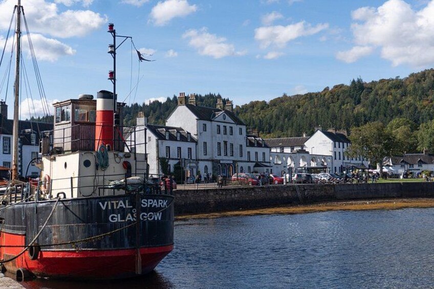 A boat named 'VITAL SPARK' docked at a harbor with buildings and a forested hill in the background. 2