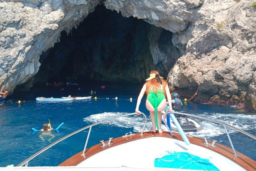 A person in a green swimsuit standing on a boat, a rocky cave entrance, and people swimming in the water. 5