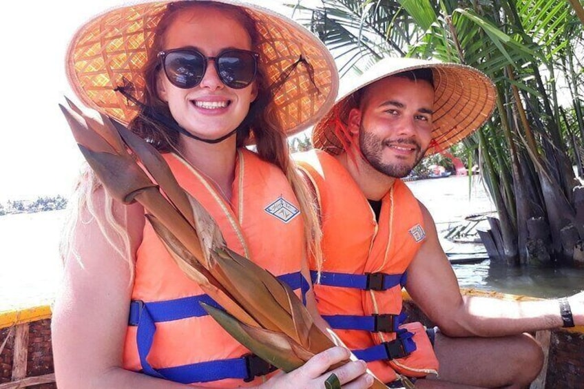 Two people wearing orange life jackets and large hats, holding a long leaf. 2