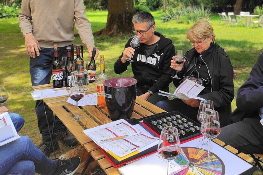 People sitting at an outdoor table with wine bottles and glasses, surrounded by papers and a wheel chart. 1