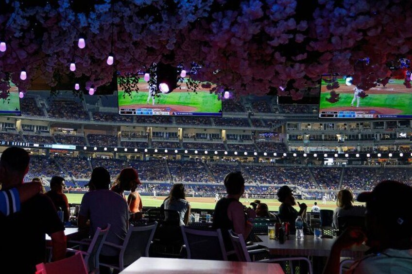 A baseball stadium with spectators seated in the stands, a large screen displaying the game, and cherry blossom trees overhead. 3