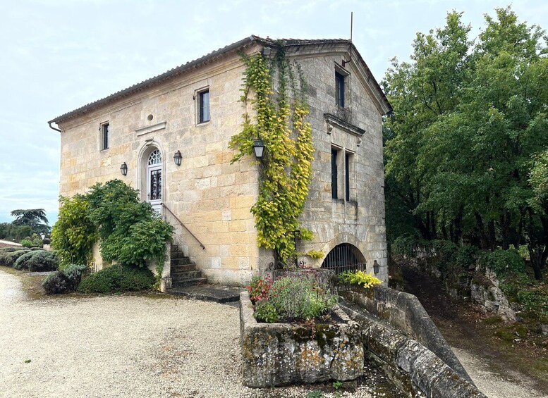 A stone building with a sloped roof, a small door, and a window. There is a staircase leading up to the entrance, and a garden with various plants and flowers. 1