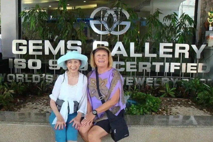 Dos personas sentadas frente a un edificio con un letrero que dice 'GEMS GALLERY ISO 9001 CERTIFIED'. 1