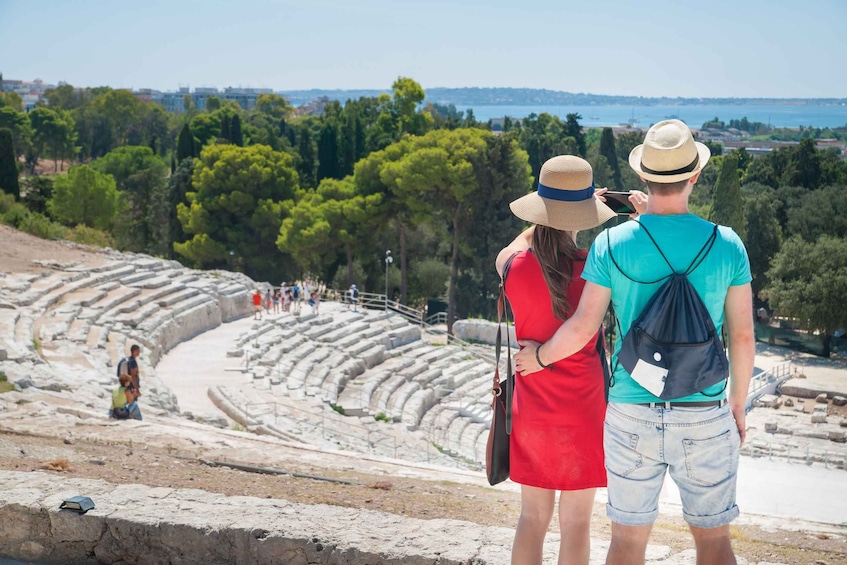 Ein Paar steht vor einem antiken Amphitheater mit Blick auf das Meer und Bäume im Hintergrund. 1
