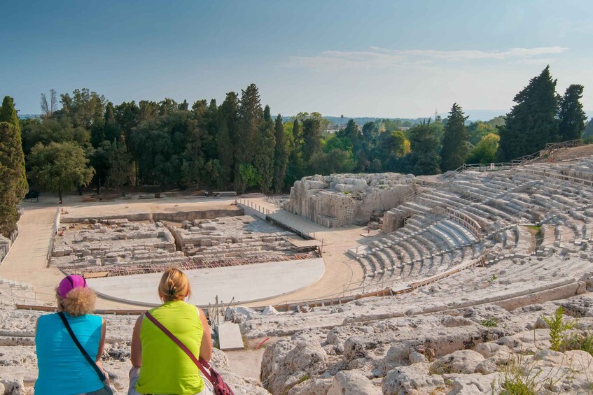 Ein antikes Amphitheater mit Steinsitzen und einer Bühne, umgeben von Bäumen und Hügeln. 2