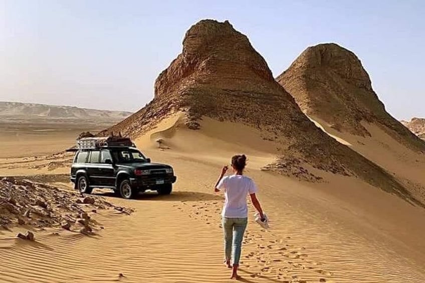 Una persona cammina verso una duna in un paesaggio desertico, con un veicolo parcheggiato nelle vicinanze. 1