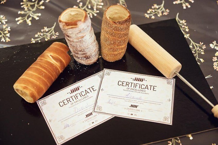 Two certificates of excellence, a rolled pastry, and a wooden rolling pin on a black surface with floral patterns. 2