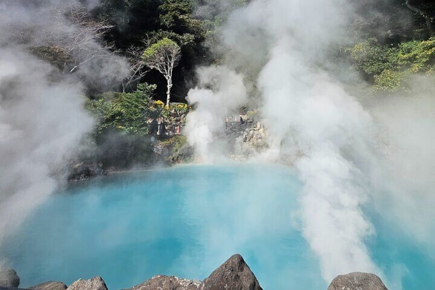 A geothermal hot spring with clear blue water, surrounded by rocks and steam rising from the ground. 1