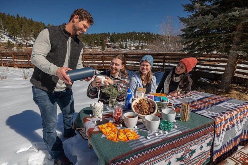 A group of people outdoors with a table set for a meal, snow on the ground, and trees in the background. 2