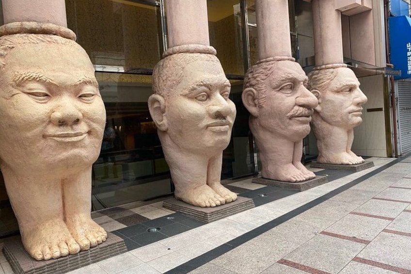 Una fila di grandi sculture in pietra con tratti umani, disposte su un pavimento piastrellato di fronte a un edificio con ampie finestre. 1