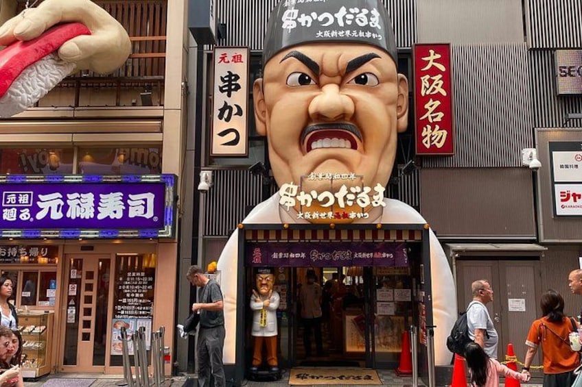 Una scena di strada con un'ampia statua di un uomo dall'espressione corrucciata, un cartello per un ristorante e un edificio con un'insegna rossa. 4