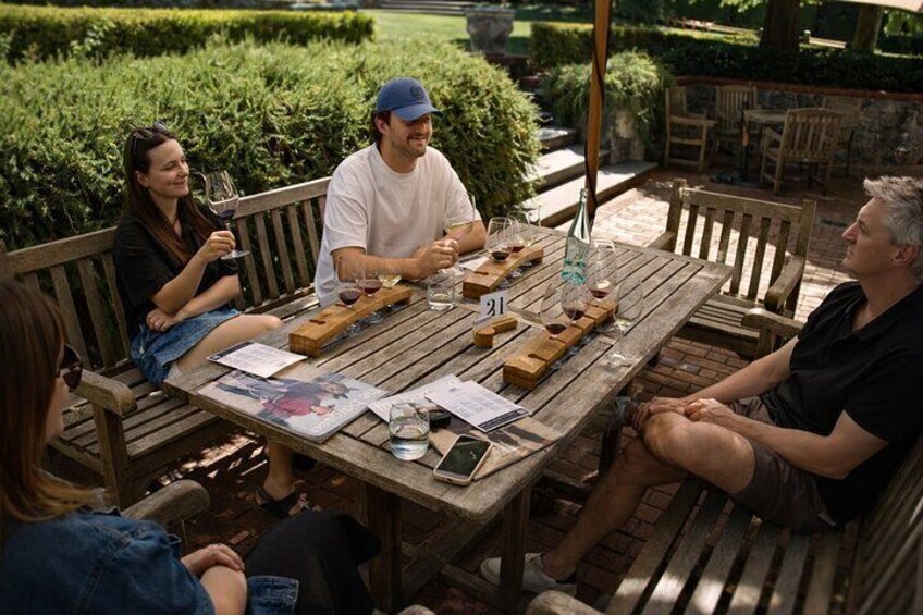 A group of people sitting around a wooden table outdoors, enjoying wine and food. 1