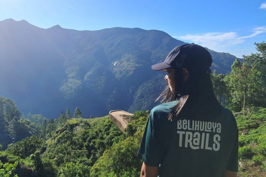 A person wearing a cap and a t-shirt with 'BELIHULAYA TRAILS' text, standing on a hill overlooking a mountainous landscape. 4