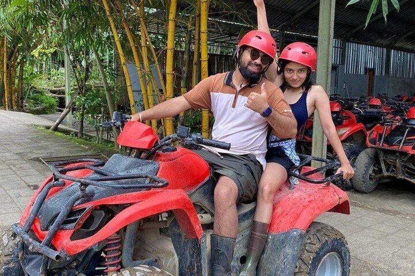Two people on a red ATV with helmets, surrounded by trees and other ATVs. 3