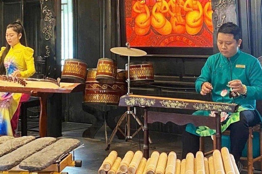 A person playing a stringed instrument, another person seated with a drum, and traditional drums on stands. 1