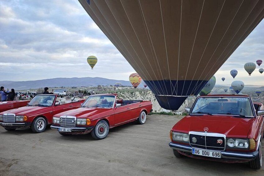 Vintage red cars parked under a large structure with hot air balloons in the background. 5