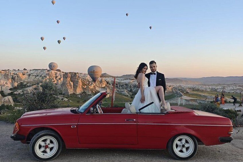 A red convertible with a couple seated, a mountainous landscape, and hot air balloons in the background. 1