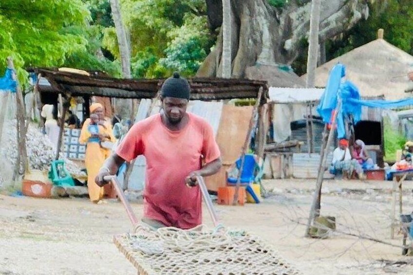 A person in a red shirt working with a woven basket, surrounded by rustic huts and trees. 3